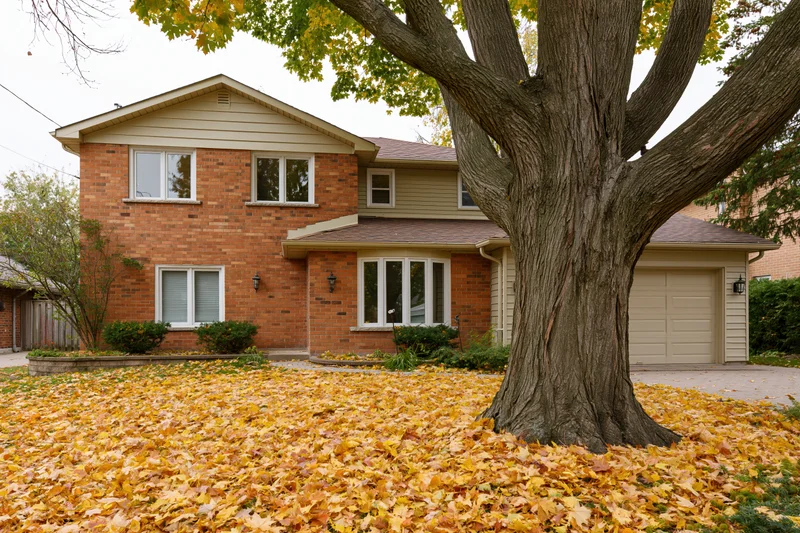 Two-story brick suburban house with beige siding and attached garage surrounded by fallen autumn leaves in front yard