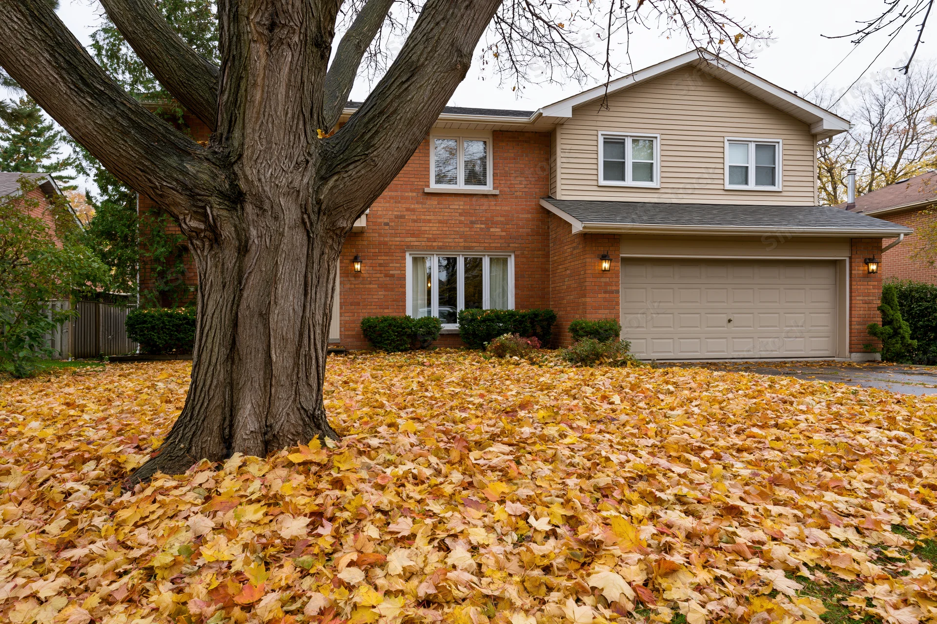 Two-story brick suburban house with beige siding and attached garage surrounded by fallen autumn leaves in front yard