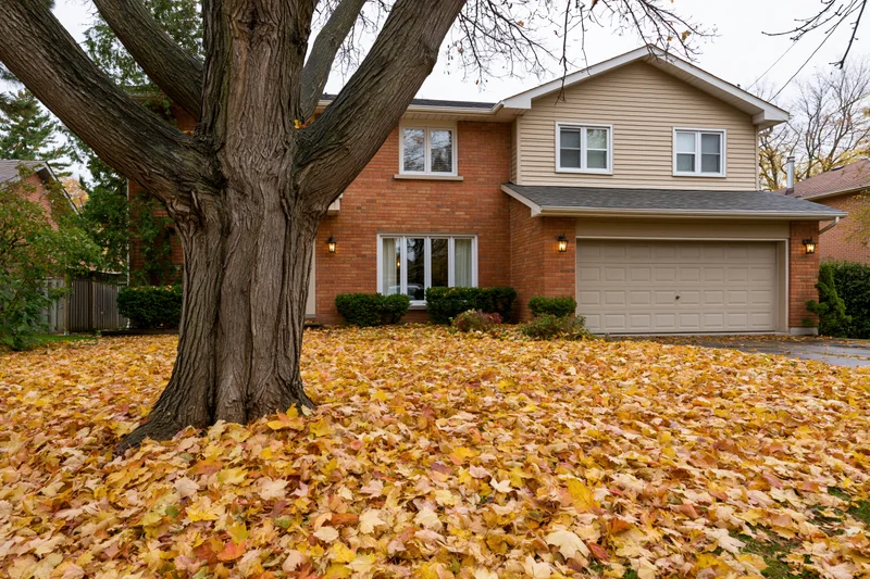 Two-story brick suburban house with beige siding and attached garage surrounded by fallen autumn leaves in front yard