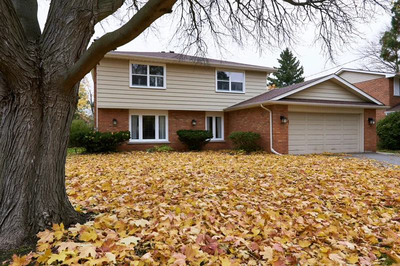 Two-story suburban house with beige siding and brick foundation surrounded by fallen autumn leaves in front yard