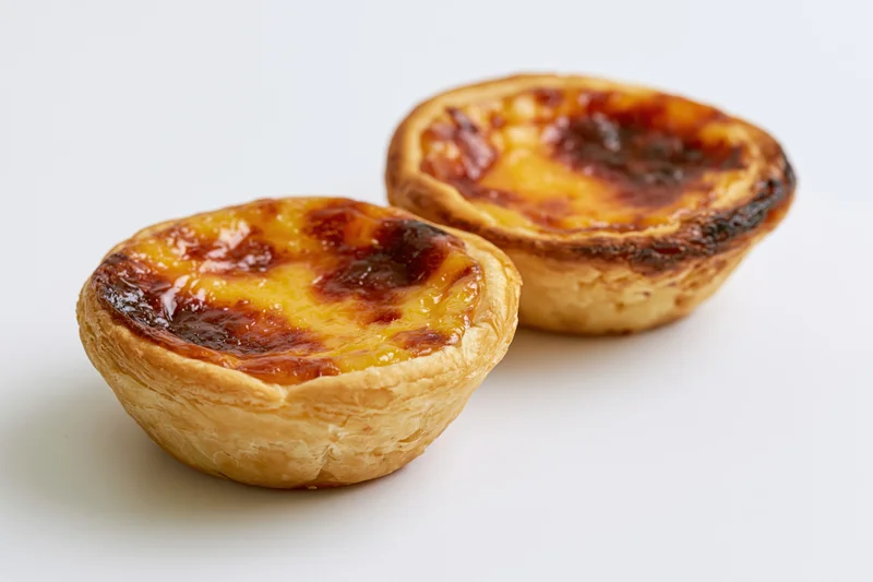 Two portuguese pastel de nata egg tarts placed close together on white surface