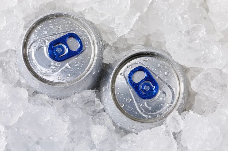 Two aluminum beverage cans with blue pull tabs resting in crushed ice