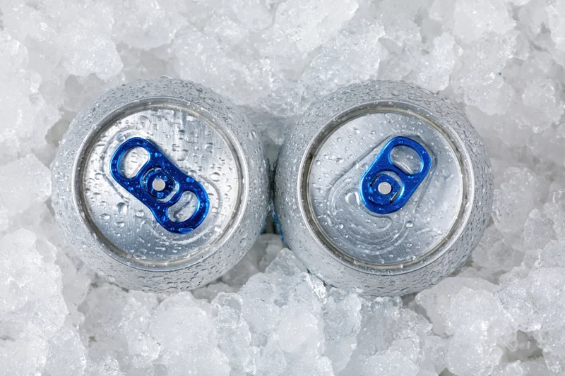 Two aluminum beverage cans with blue pull tabs resting in crushed ice