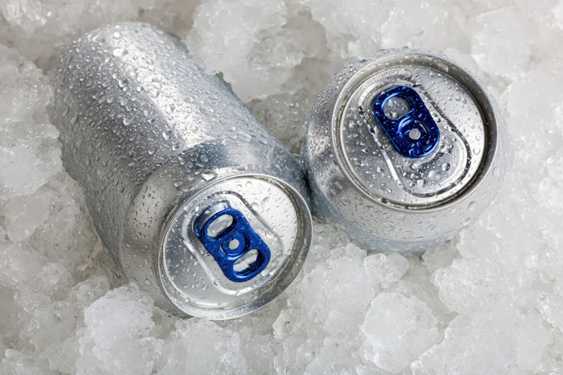 Two aluminum beverage cans with blue pull tabs resting in crushed ice
