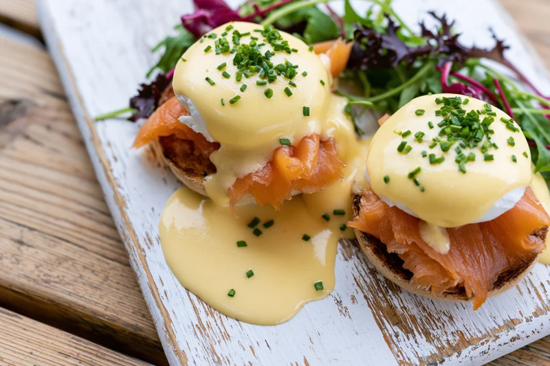 Two eggs benedict with smoked salmon on toasted english muffins served on a white wooden board with mixed greens and hollandaise sauce