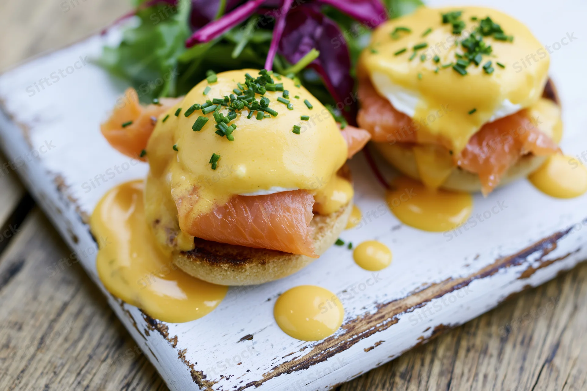 Two eggs benedict with smoked salmon on toasted english muffins served with mixed greens on a rustic white plate