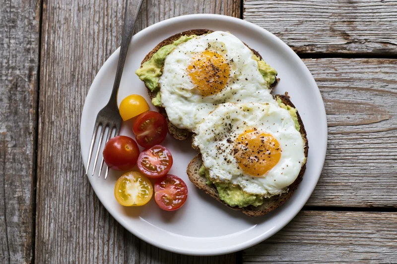 Two fried eggs on avocado toast with cherry tomatoes on a white plate on wooden table