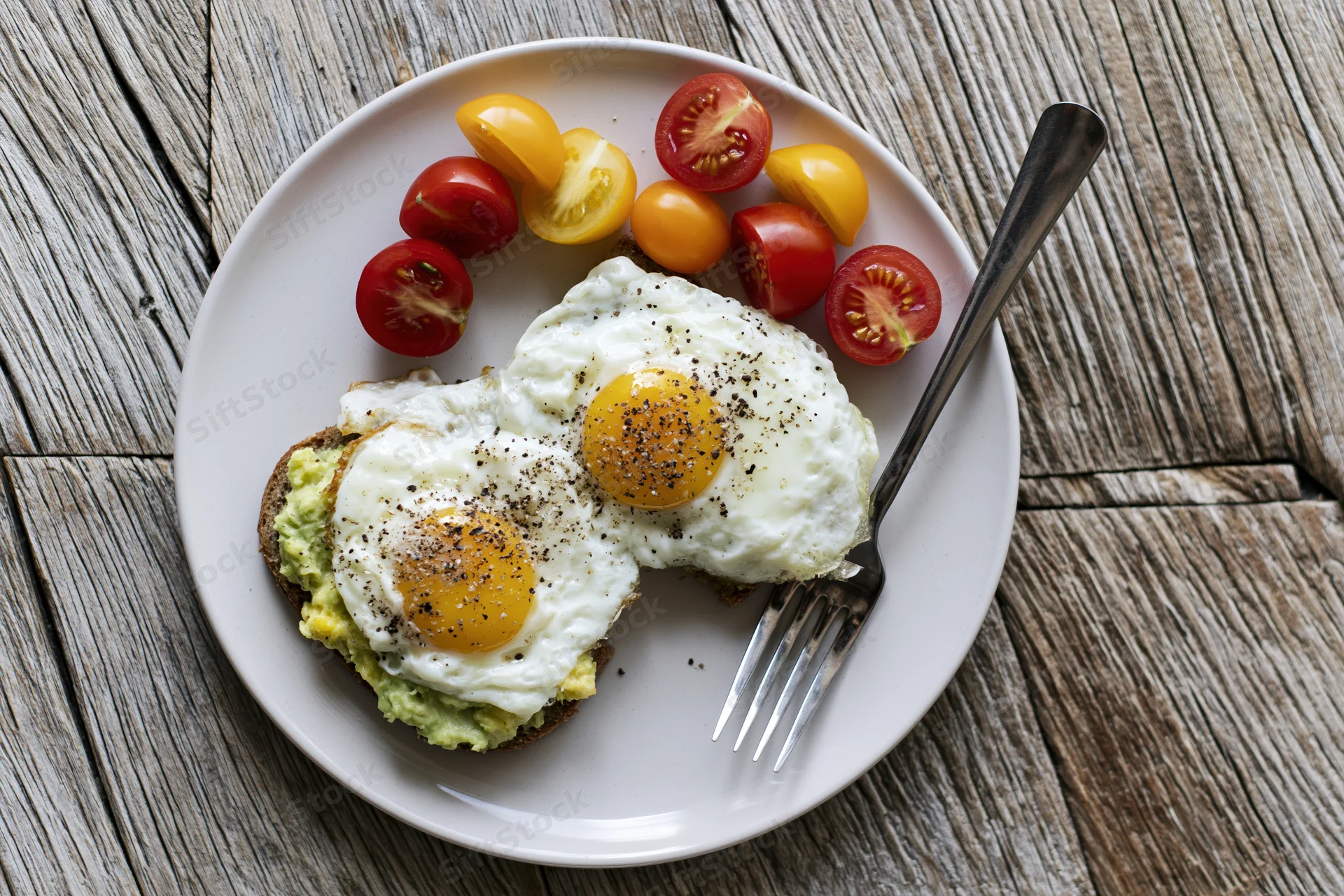 Two fried eggs on avocado toast with cherry tomatoes on a white plate on wooden table