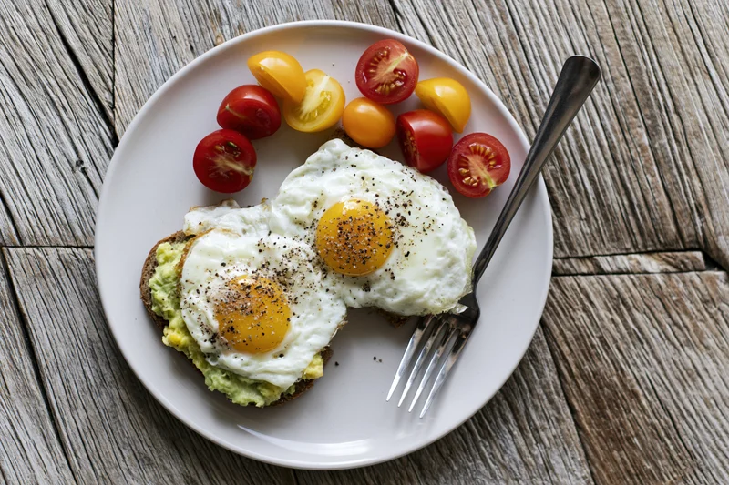 Two fried eggs on avocado toast with cherry tomatoes on a white plate on wooden table