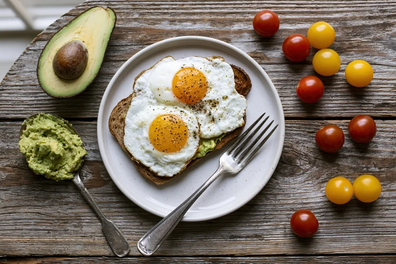 Two fried eggs on avocado toast served on a white ceramic plate with cherry tomatoes and avocado on a rustic wooden table
