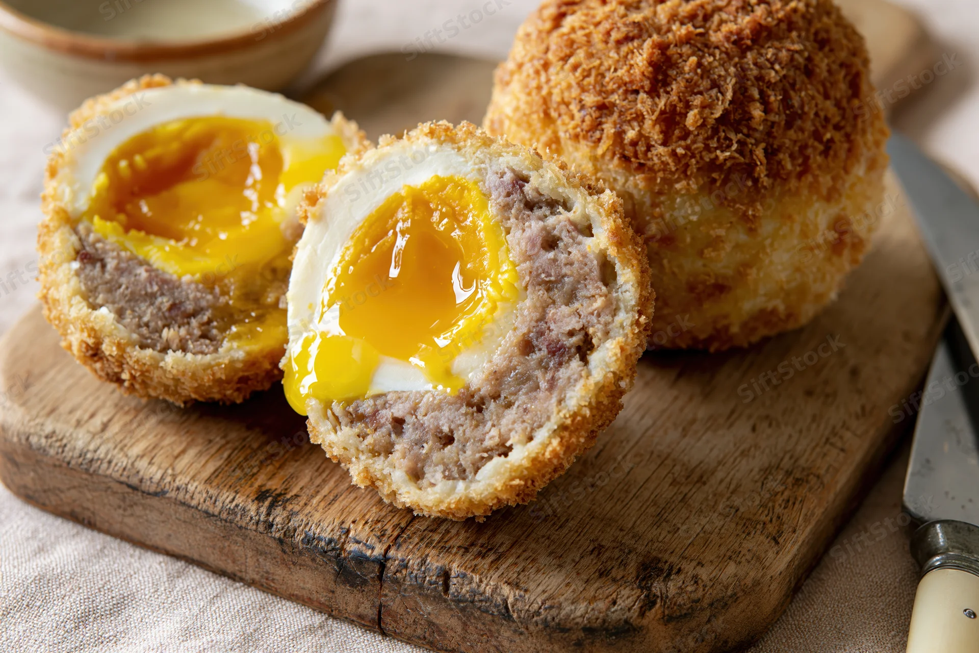 Halved scotch eggs with runny yolks on a wooden cutting board beside a knife