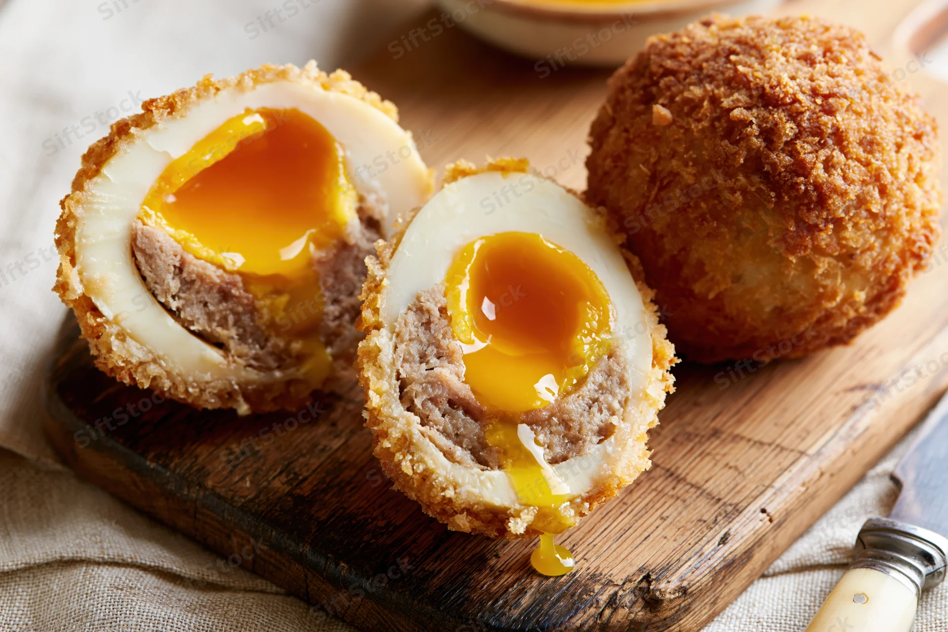 Halved scotch eggs with runny yolk on wooden cutting board beside whole egg and knife