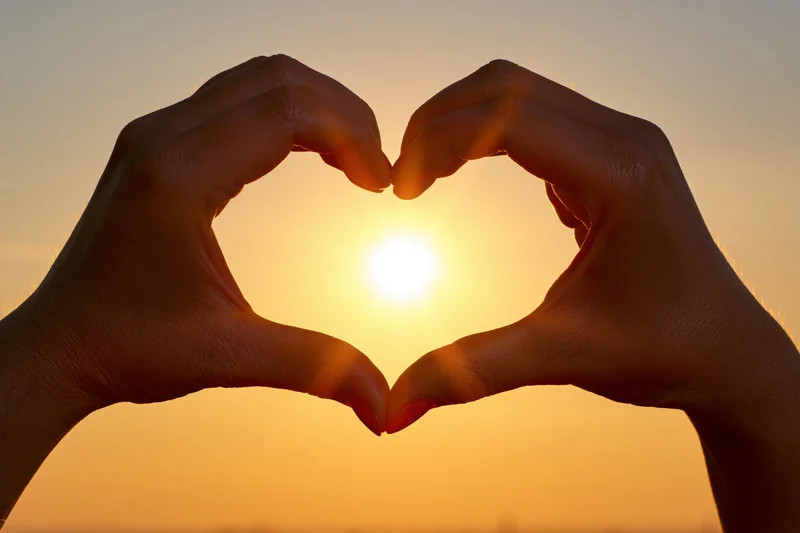 Hands forming heart shape silhouetted against bright sunset sky