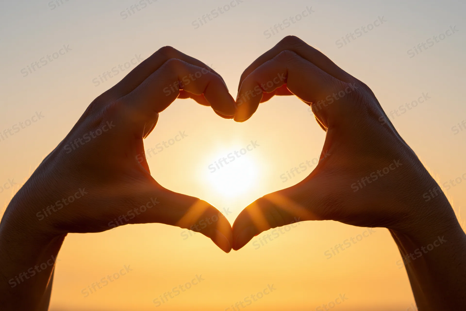 Hands forming heart shape silhouetted against bright sunset sky