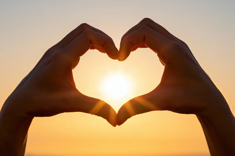 Hands forming heart shape silhouetted against bright sunset sky