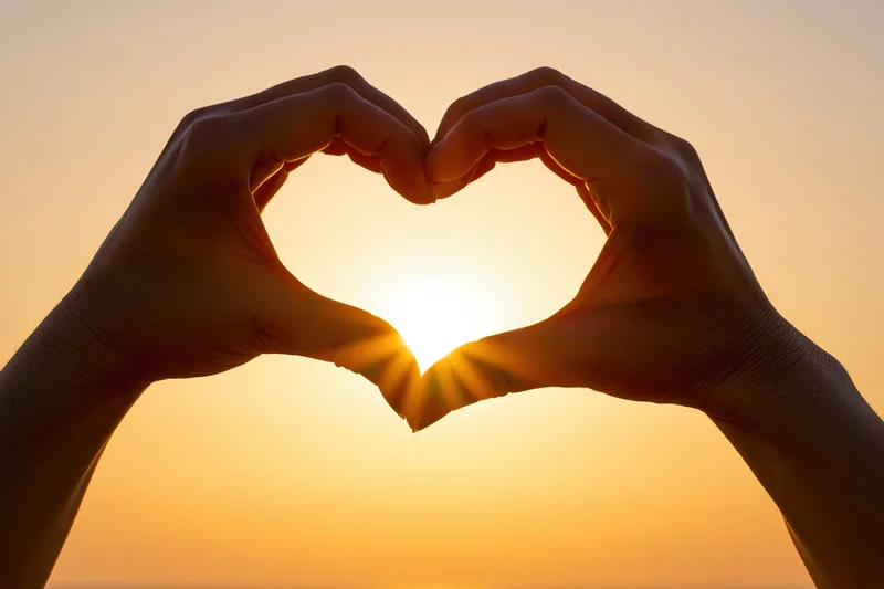 Hands forming heart shape silhouetted against bright sunset sky