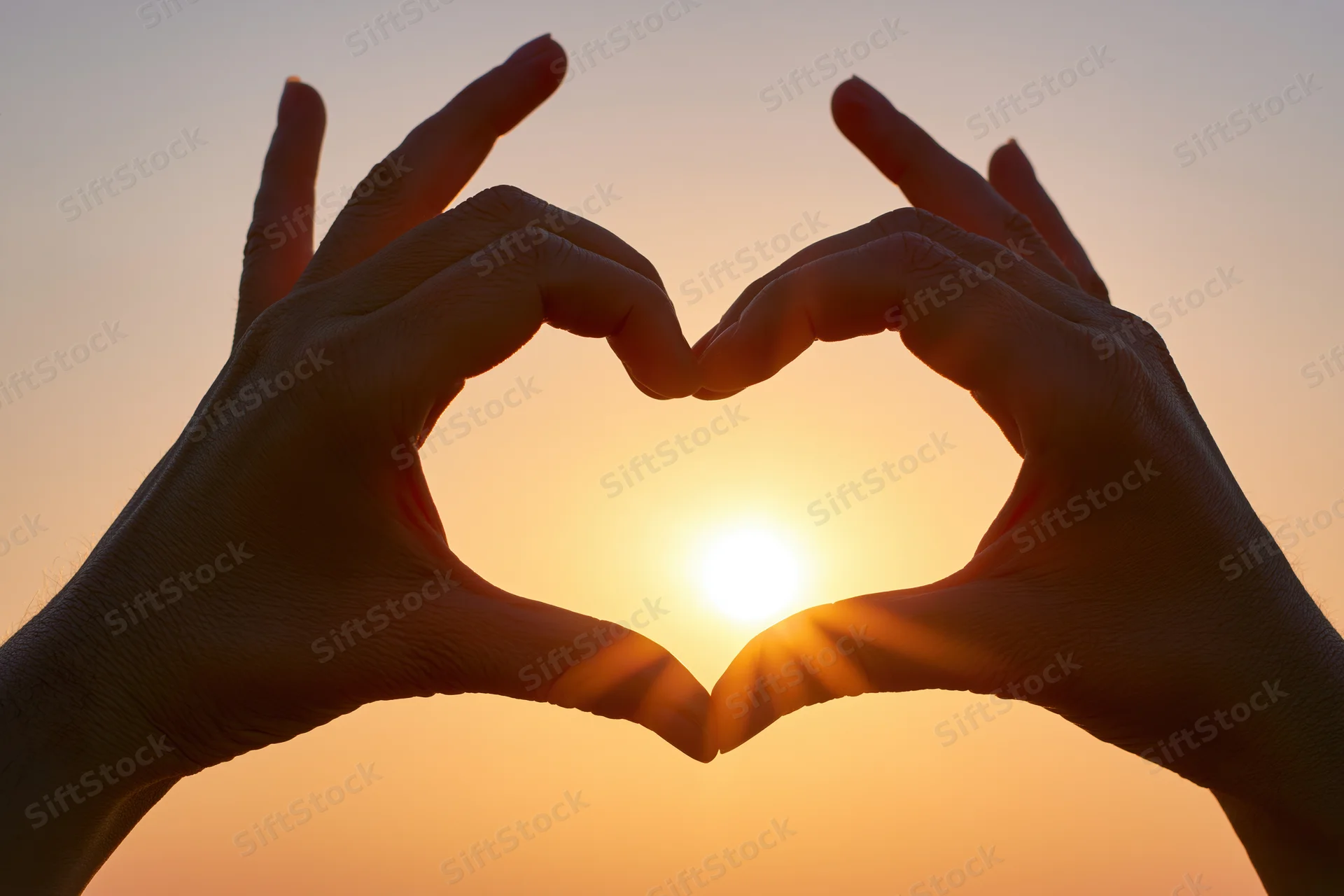 Hands forming a heart shape silhouetted against a bright sunset sky