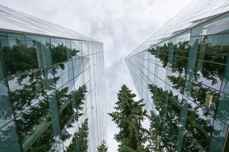 Two modern glass office buildings viewed from ground level with reflections of evergreen trees and cloudy sky