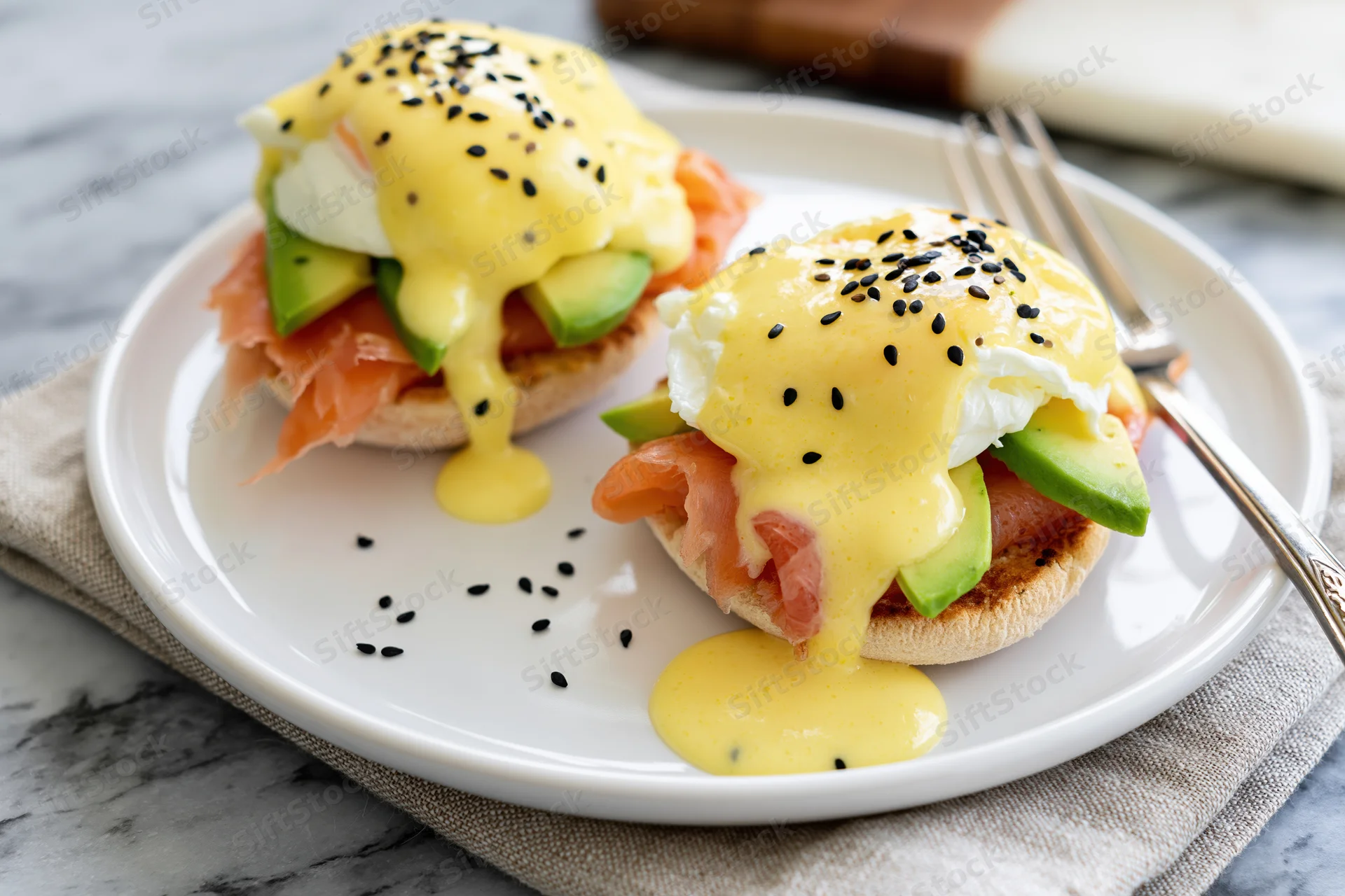 Two open-faced egg benedict toasts with salmon and avocado on a white ceramic plate with hollandaise sauce and black sesame seeds, served on a marble table with a fork and napkin