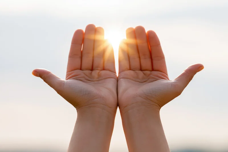 Open hands raised upward toward bright glowing sun in sky