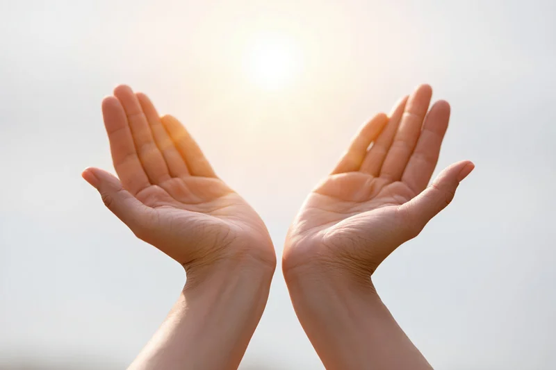 Open cupped hands raised upward toward a bright glowing sun in the sky