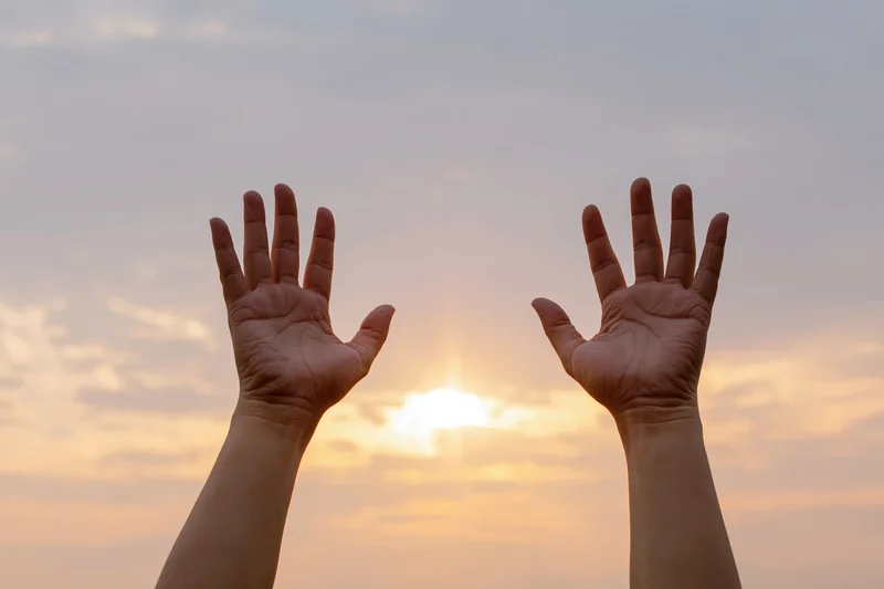 Two open hands raised upward against a wide sunset sky framing the sun