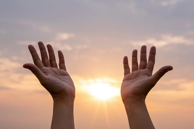 Two open hands raised upward against a wide sunset sky framing the sun