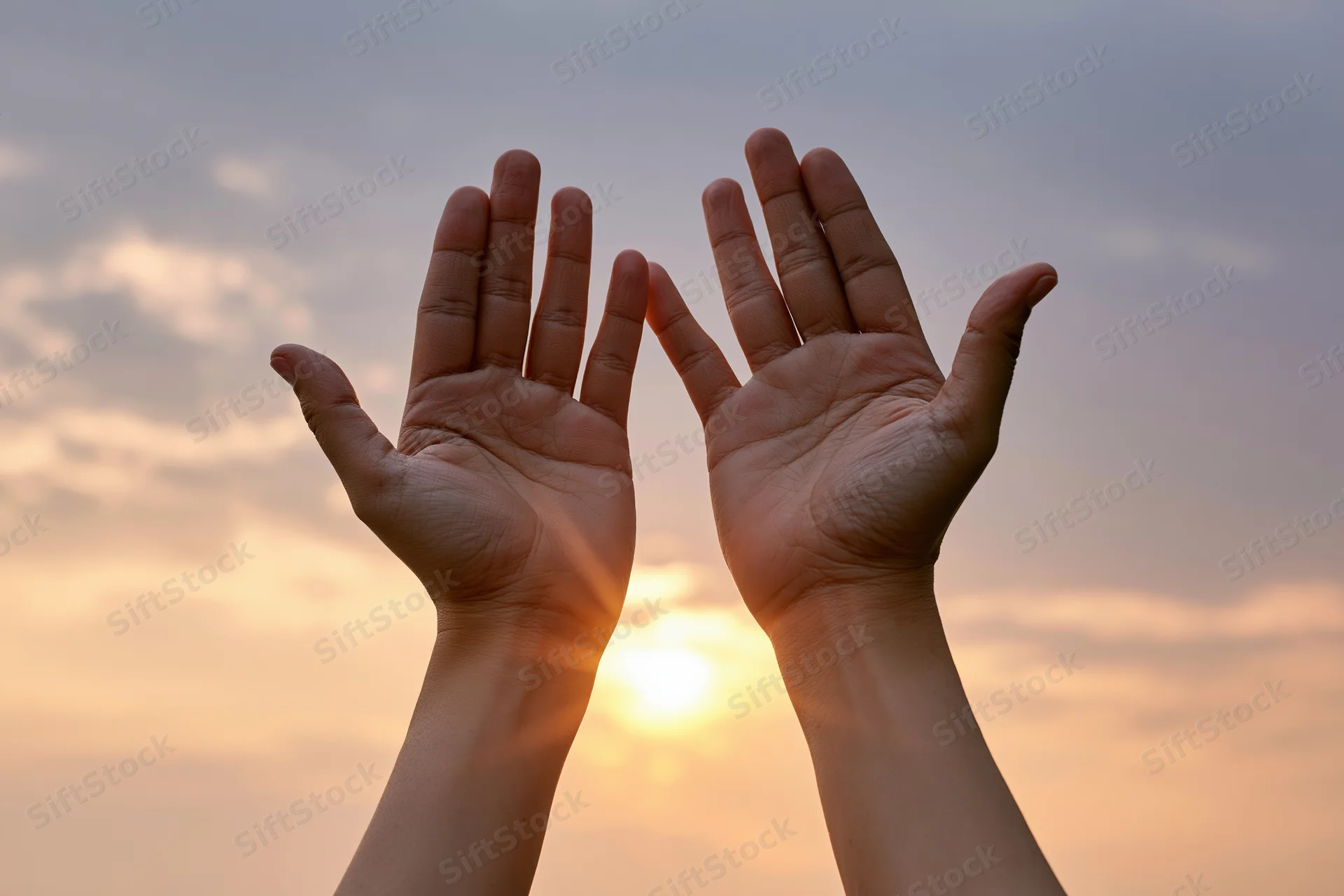 Two open hands raised upward against a wide sunset sky framing the sun