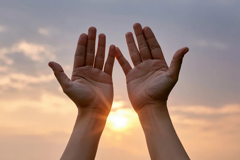 Two open hands raised upward against a wide sunset sky framing the sun