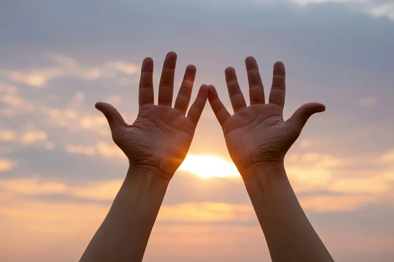 Two open hands raised upward framing the sun against a colorful sunset sky