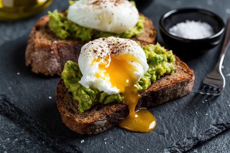 Two slices of sourdough toast topped with smashed avocado and poached eggs, one with runny yolk, on a dark slate surface with salt and oil nearby