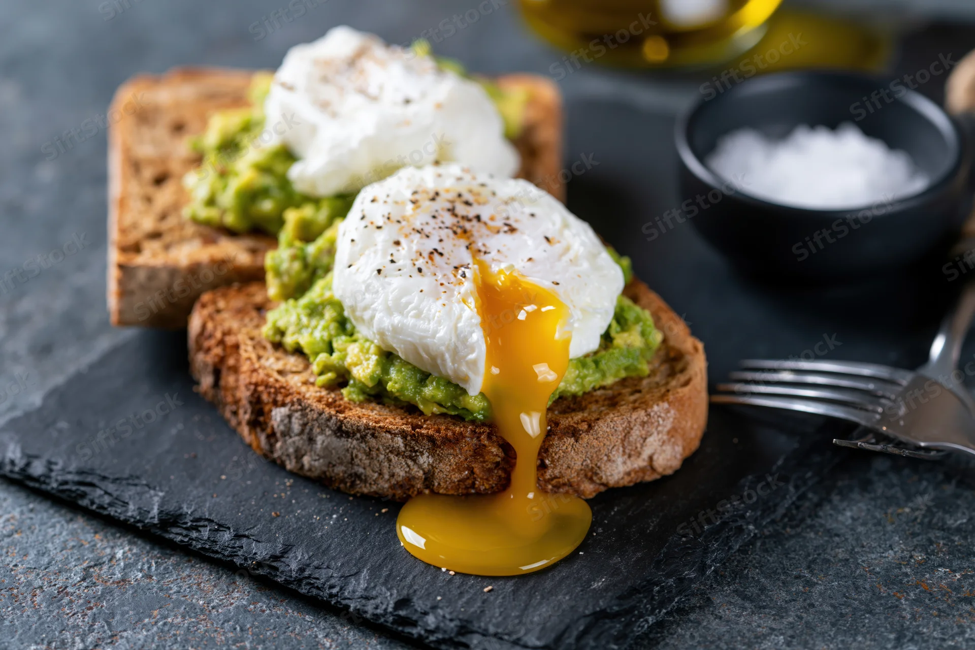 Two slices of sourdough toast with smashed avocado and poached eggs, one with runny yolk dripping, on a slate plate with salt and olive oil nearby