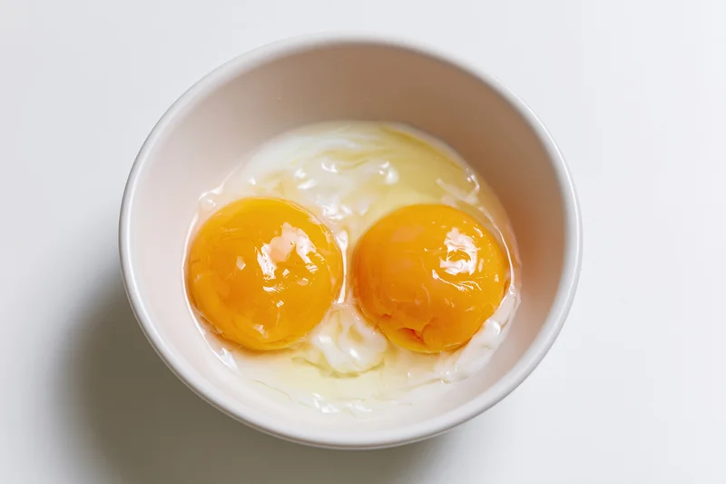 Two raw cracked eggs with bright orange-yellow yolks in a white ceramic bowl on a plain white surface