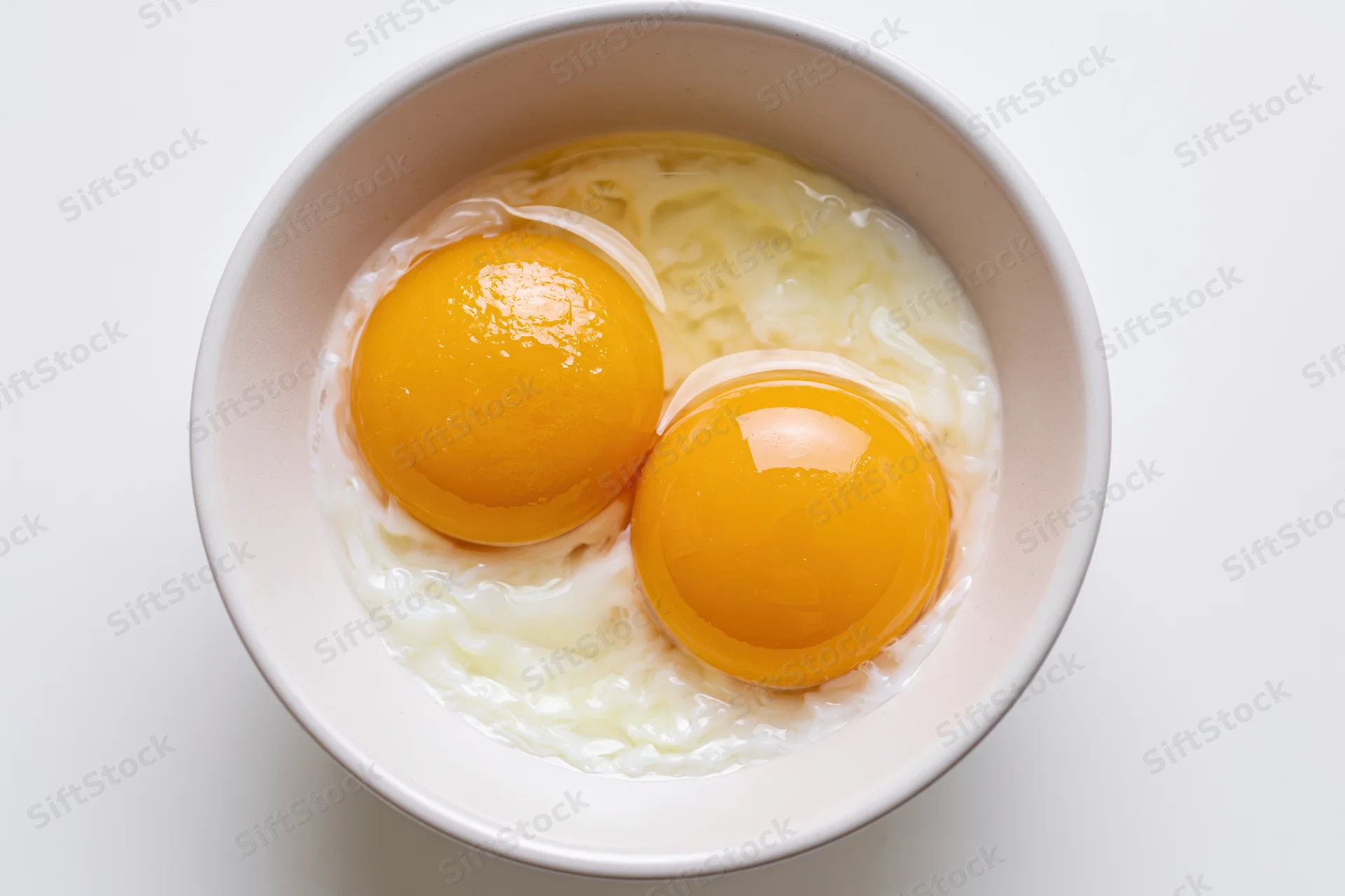 Two raw cracked eggs with bright orange-yellow yolks in a white bowl on a plain surface