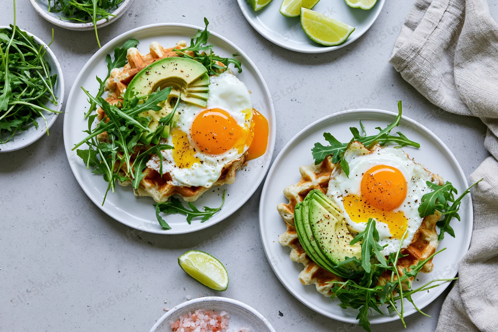 Two savory waffles topped with sliced avocado and fried eggs, garnished with arugula and lime wedges on white plates on a light gray surface