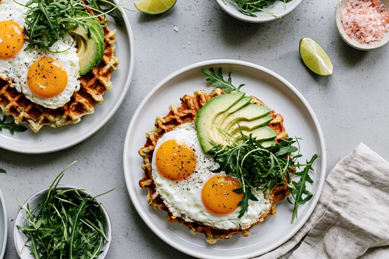 Two savory waffles topped with fried eggs, sliced avocado, and arugula on white plates with lime and salt on a gray surface