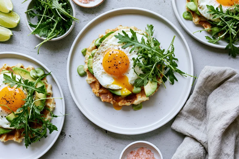 Savory waffles topped with fried eggs avocado and arugula on white plates arranged on a table