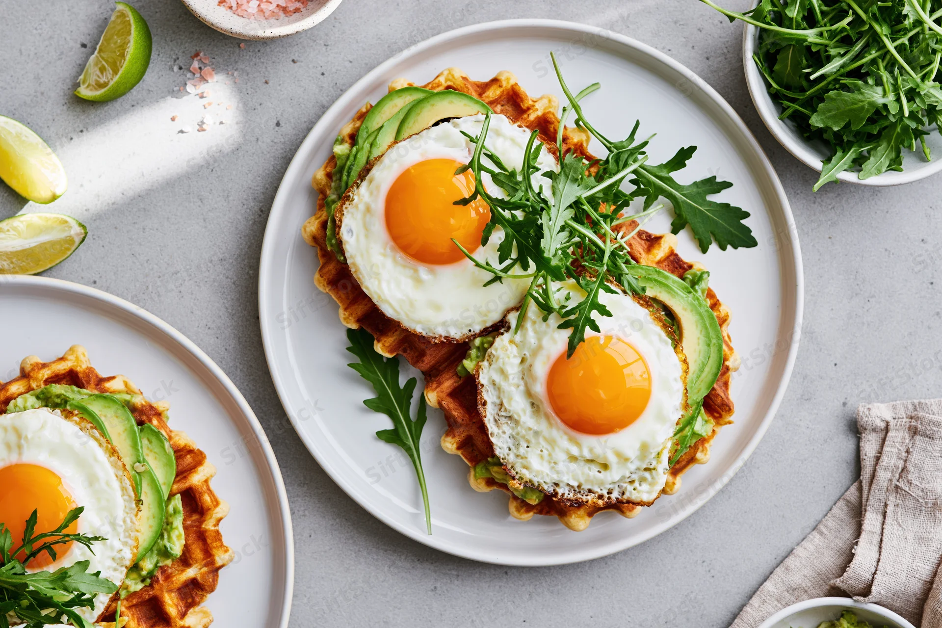 Two savory waffles topped with avocado and fried eggs on white plates with arugula and lime garnish on a gray surface