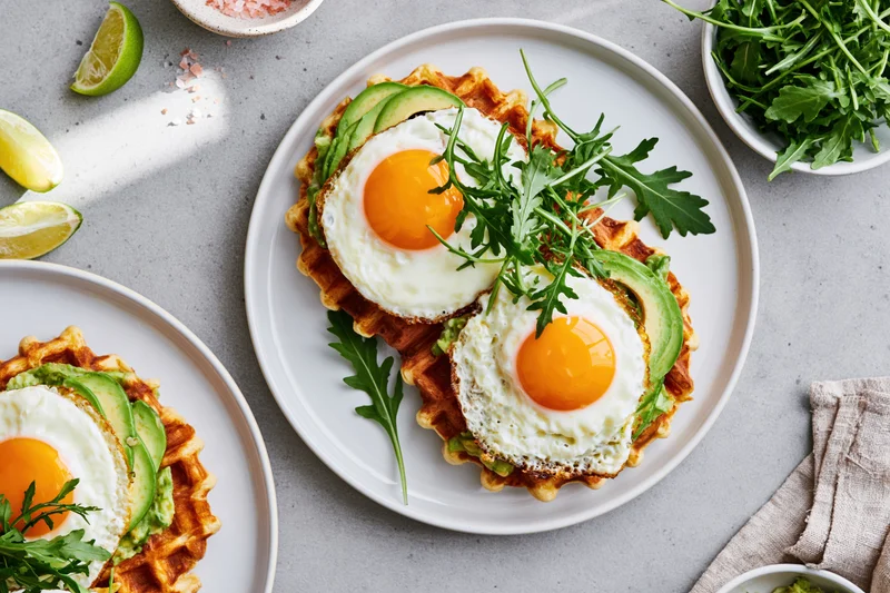 Two savory waffles topped with avocado and fried eggs on white plates with arugula and lime garnish on a gray surface