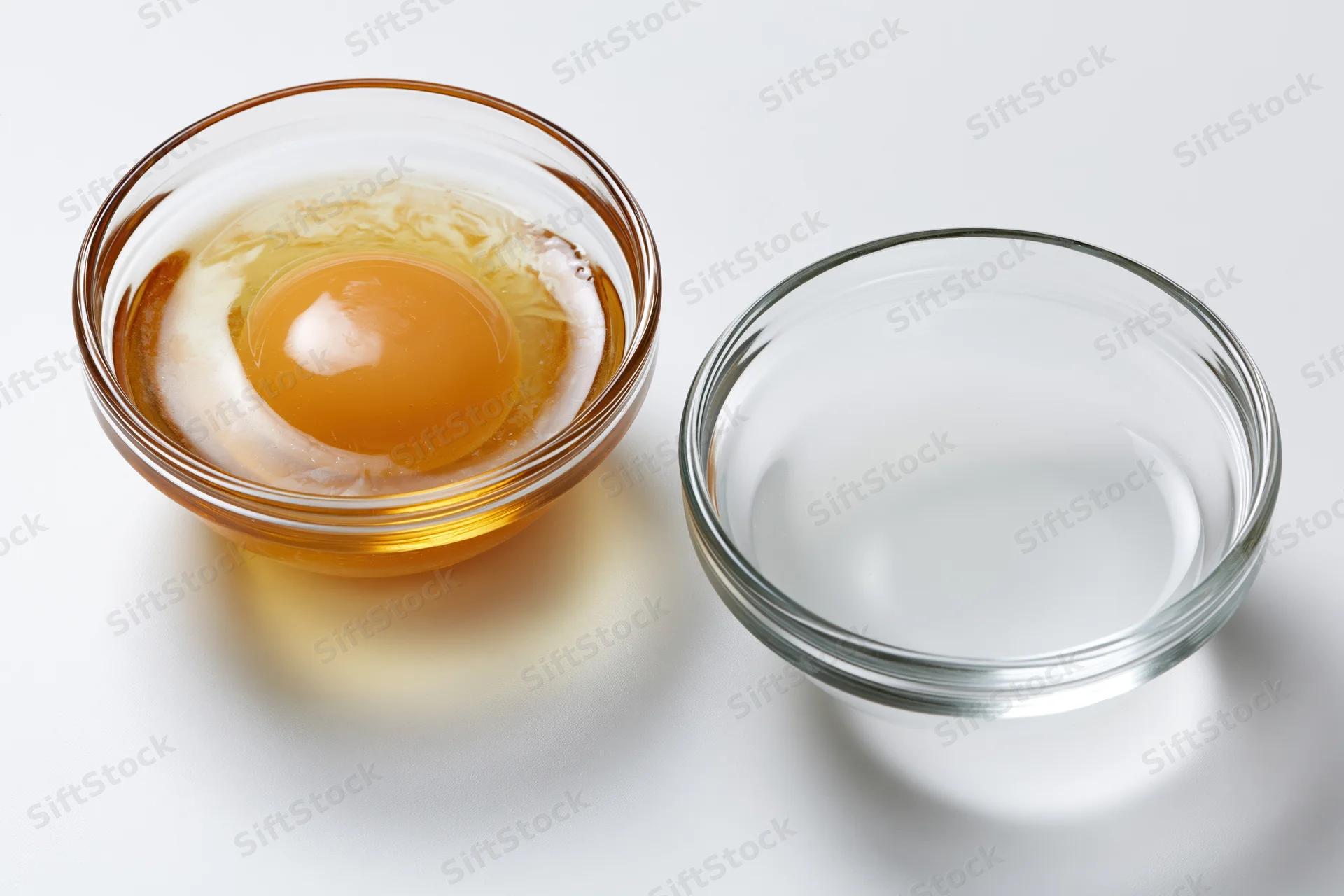 Raw egg in a clear glass bowl beside an empty one on a white surface