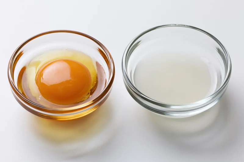 Two small clear glass bowls side by side on white surface, one containing raw egg yolk and the other egg white