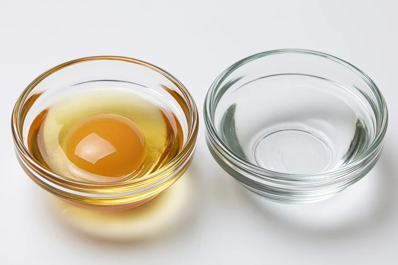 Raw egg in a small clear glass bowl beside an empty one on a white surface