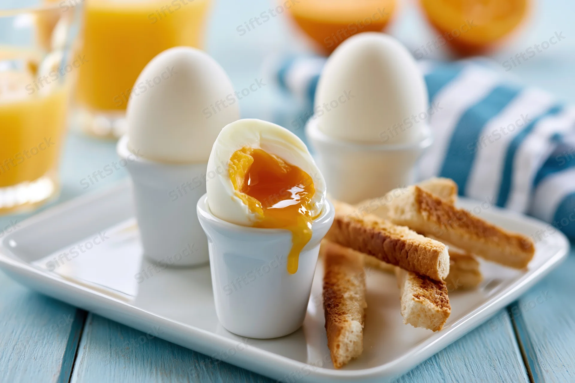 Soft-boiled eggs in white ceramic egg cups with toast and orange juice on a white plate