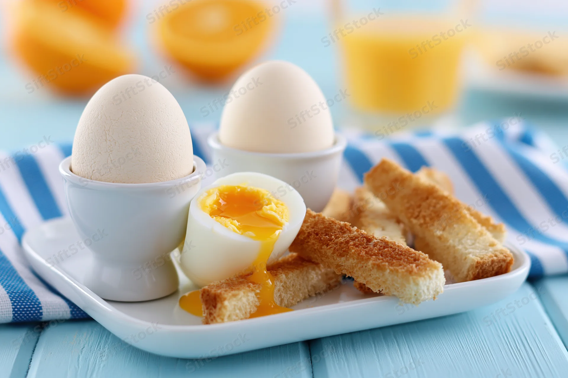 Two soft-boiled eggs in white ceramic egg cups with toast and orange juice on a blue striped cloth at breakfast