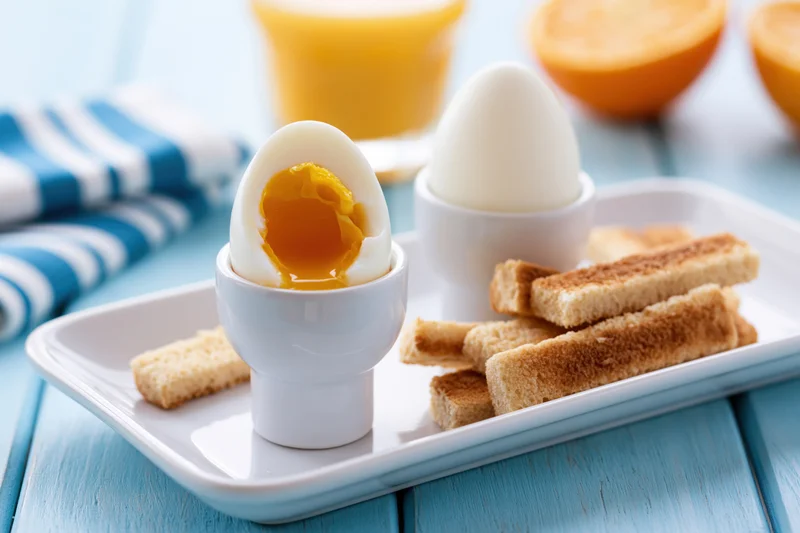 Two soft-boiled eggs in white ceramic egg cups on a white tray with toast and orange juice on a blue wooden table