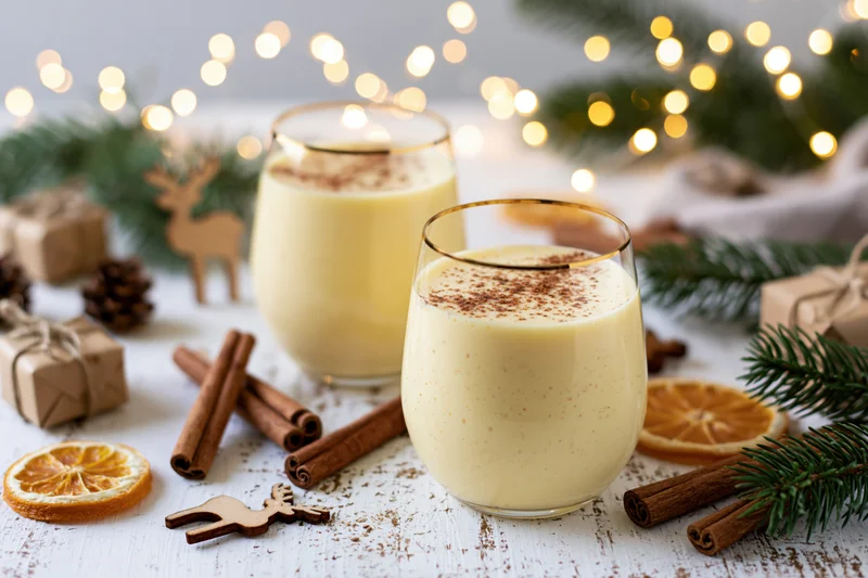 Two stemless glasses of creamy pale yellow eggnog dusted with cinnamon on a white rustic table surrounded by holiday decorations