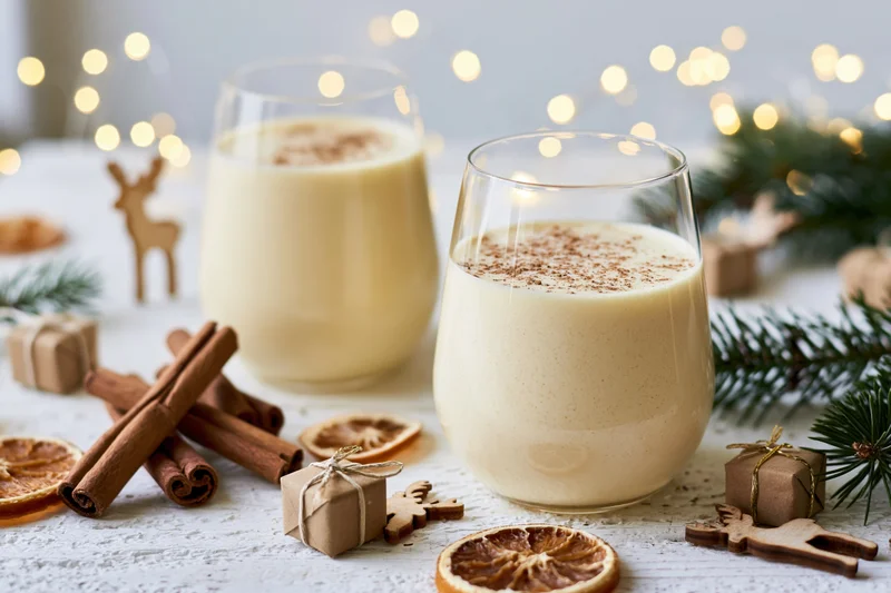 Two stemless glasses of creamy pale yellow eggnog dusted with cinnamon on a white table with holiday decorations and bokeh lights in the background