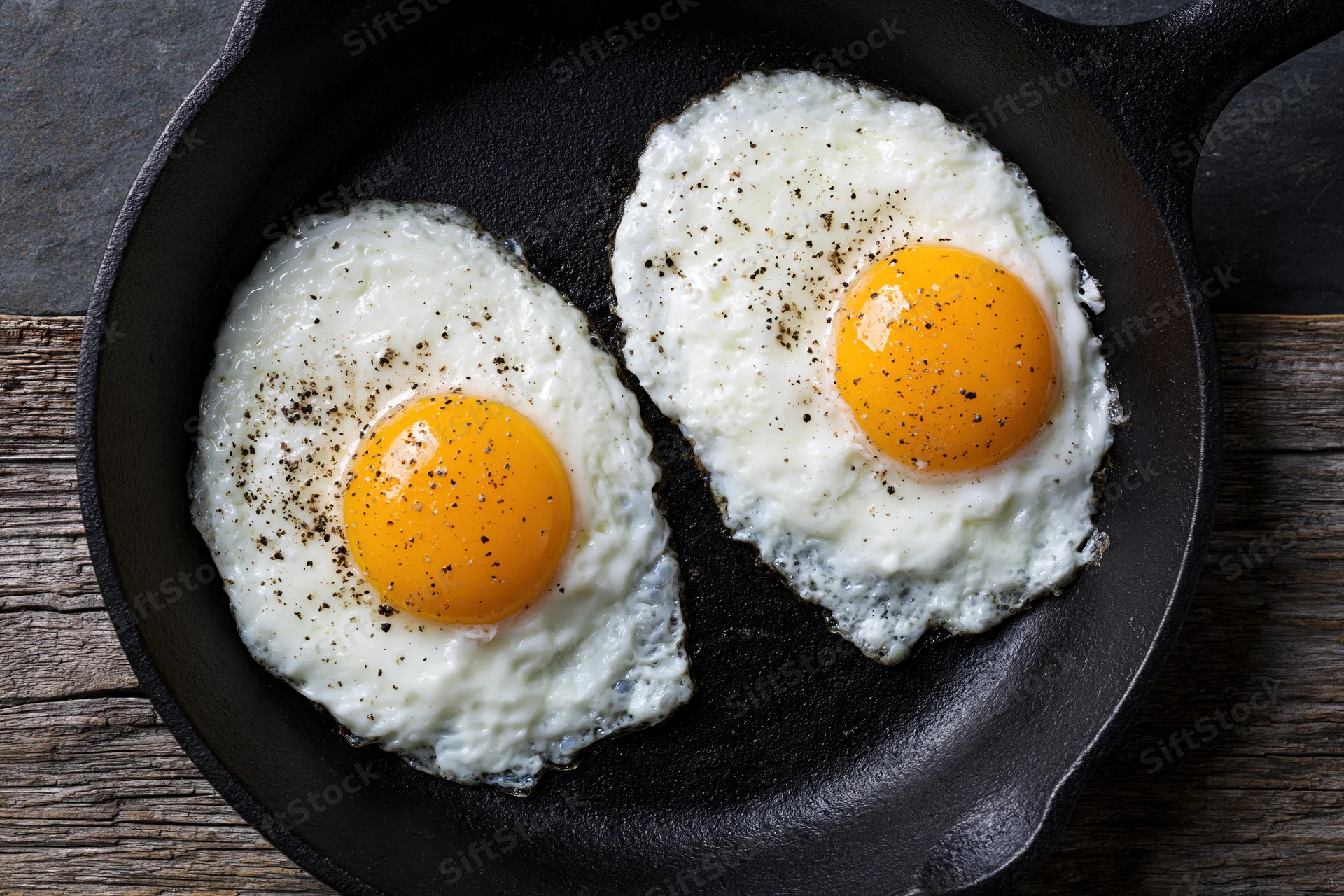 Two sunny-side-up fried eggs with bright orange yolks in a cast iron skillet on a wooden surface