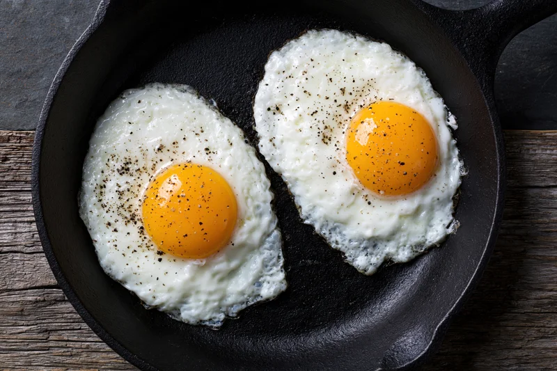 Two sunny-side-up fried eggs with bright orange yolks in a cast iron skillet on a wooden surface