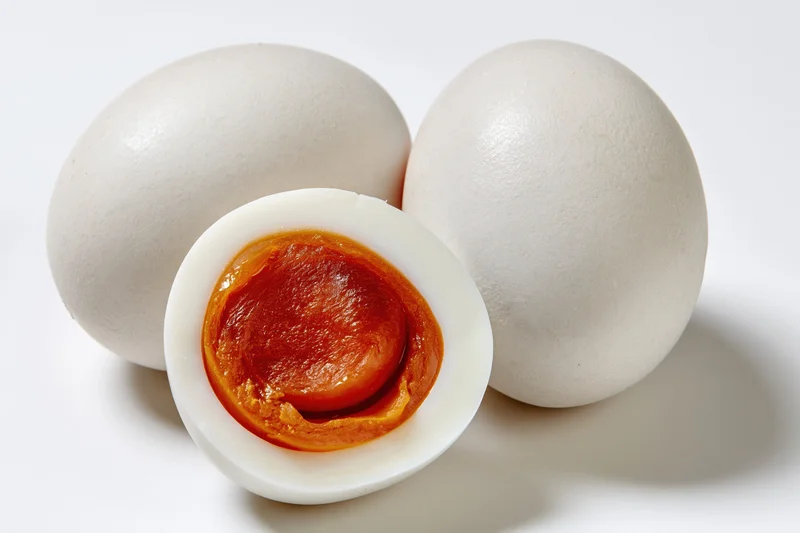 Two whole salted duck eggs and one halved egg showing orange yolk on white background